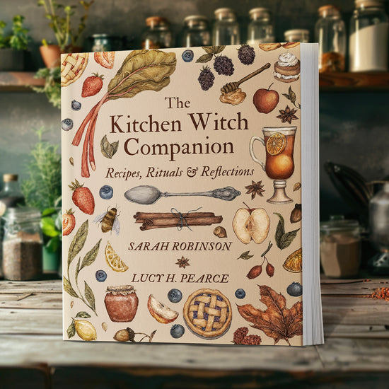 A tan softcover book standing on a wood table. In the background are glass jars willed with herbs and spices, sitting on wood shelves. On the book cover are illustrations of fruits, pies, herbs, leaves, etc. Brown text says "The Kitchen Witch Companion: Recipes, Rituals & Reflections."