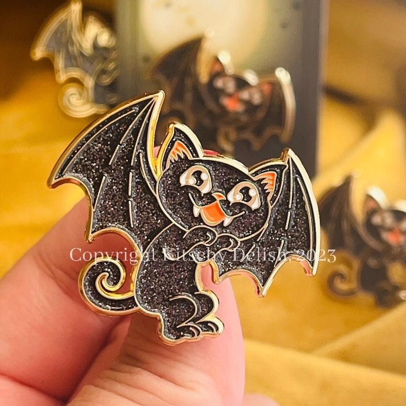 A close-up of a hand holding a black cat bat enamel pin with gold trim and orange accents, with blurred bat pins in the background.