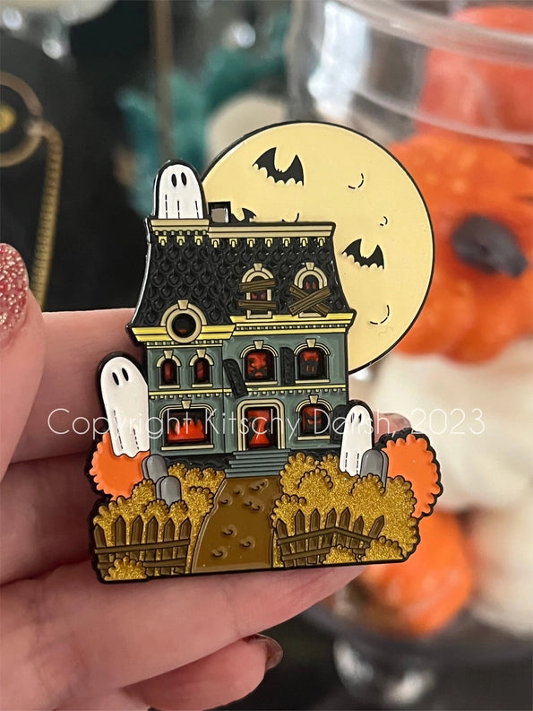 An enamel pin depicting a haunted house with a moon on the top corner