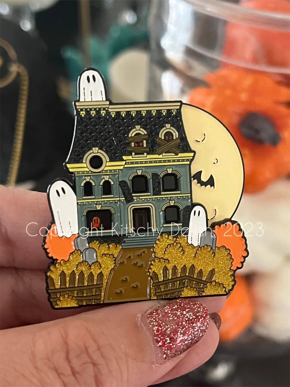 An enamel pin depicting a haunted house with a moon on the top corner