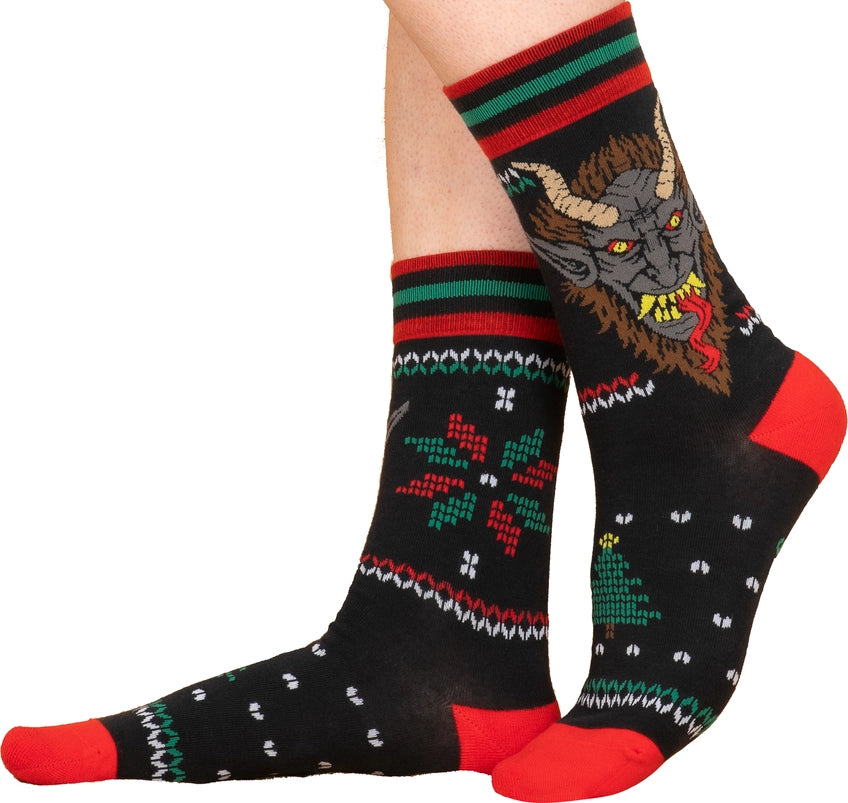 A model's feet wearing a pair of black socks with red and green stripes at the top. One side has a depiction of a red and green snowflake shape. The other side depicts the face of the mythological figure Krampus, with long horns on his head, sharp teeth, and a long red tongue.