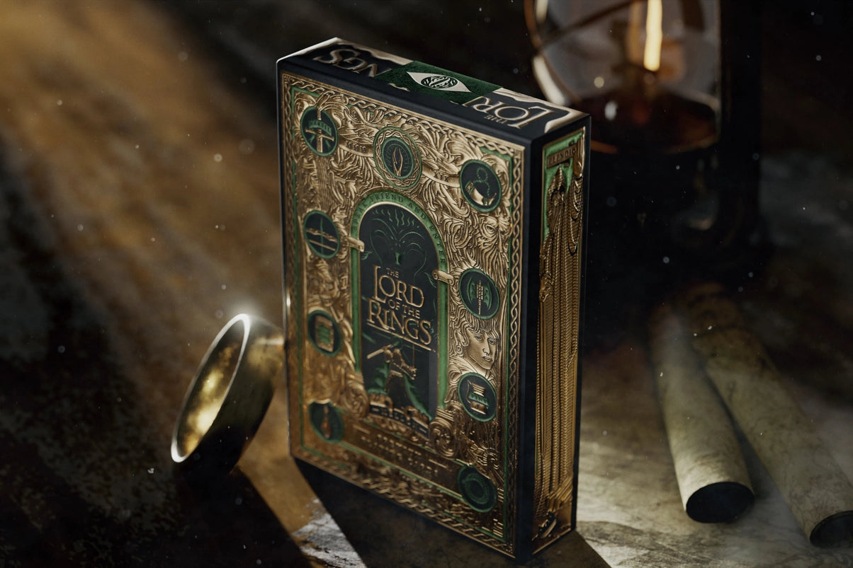 A green and gold box of playing cards, on a wooden table. The box front depicts the Lord of the Rings book cover. Next to the box is a gold ring. In the background is an oil lamp.