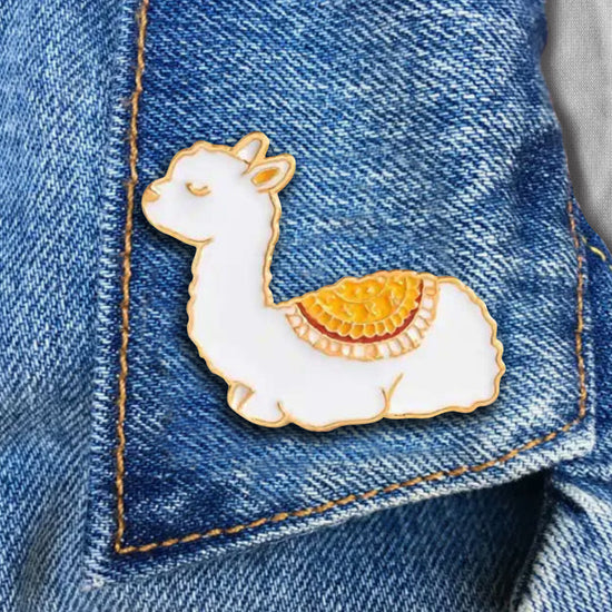 Close up of a enamel pin attached to a light blue demin collar. The pin is shaped like an alpaca sitting down. A gold and red round blanket is on the alpaca's back.