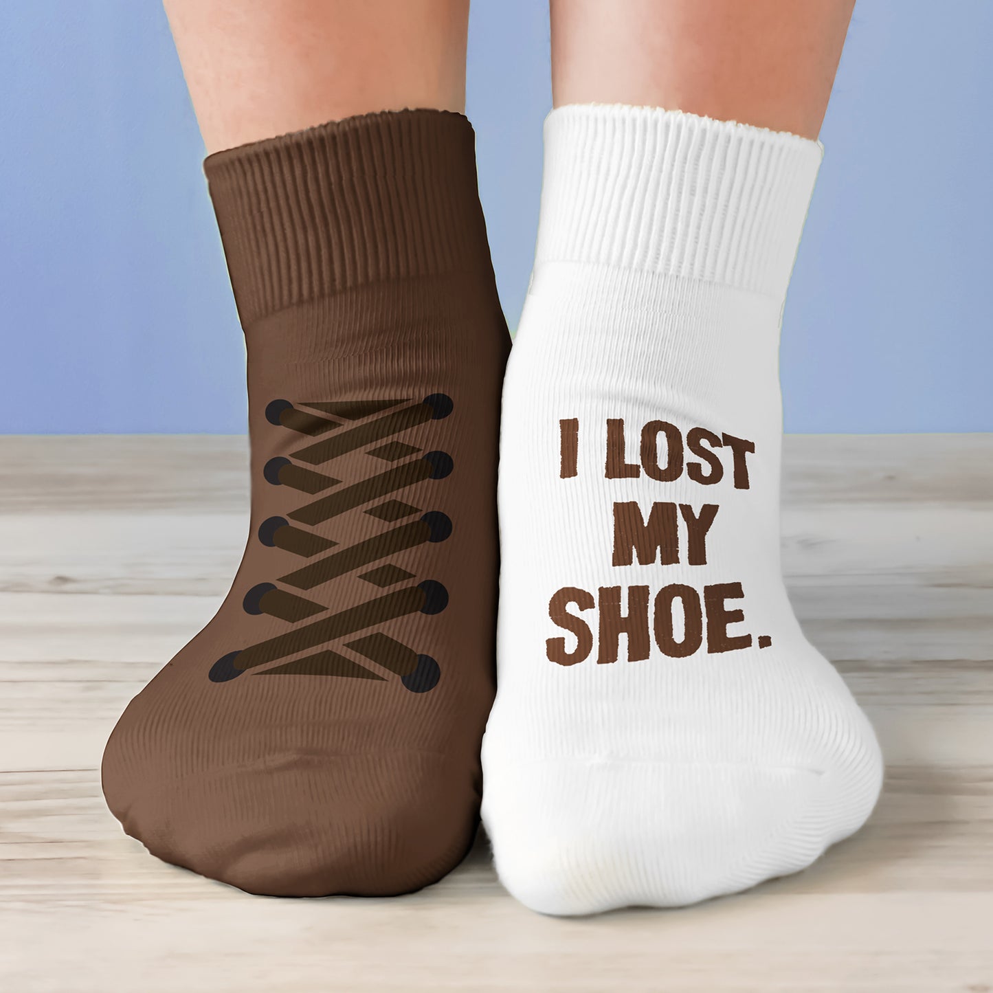 A model's feet wearing one brown sock and one white sock. The brown sock has shoelaces printed on the top. The right sock has brown text saying "I lost my shoe."