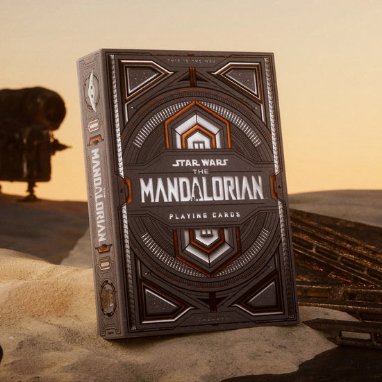 A brown and silver box of playing cards, on a patch of desert sand. The box depicts images from the TV series "The Mandalorian." In the background is a spaceship from the series.