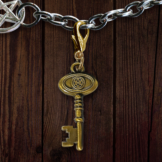 A gold-colored skeleton key on a silver chain, against a wooden background. The key depicts the Men Of Letters symbol at the bow.