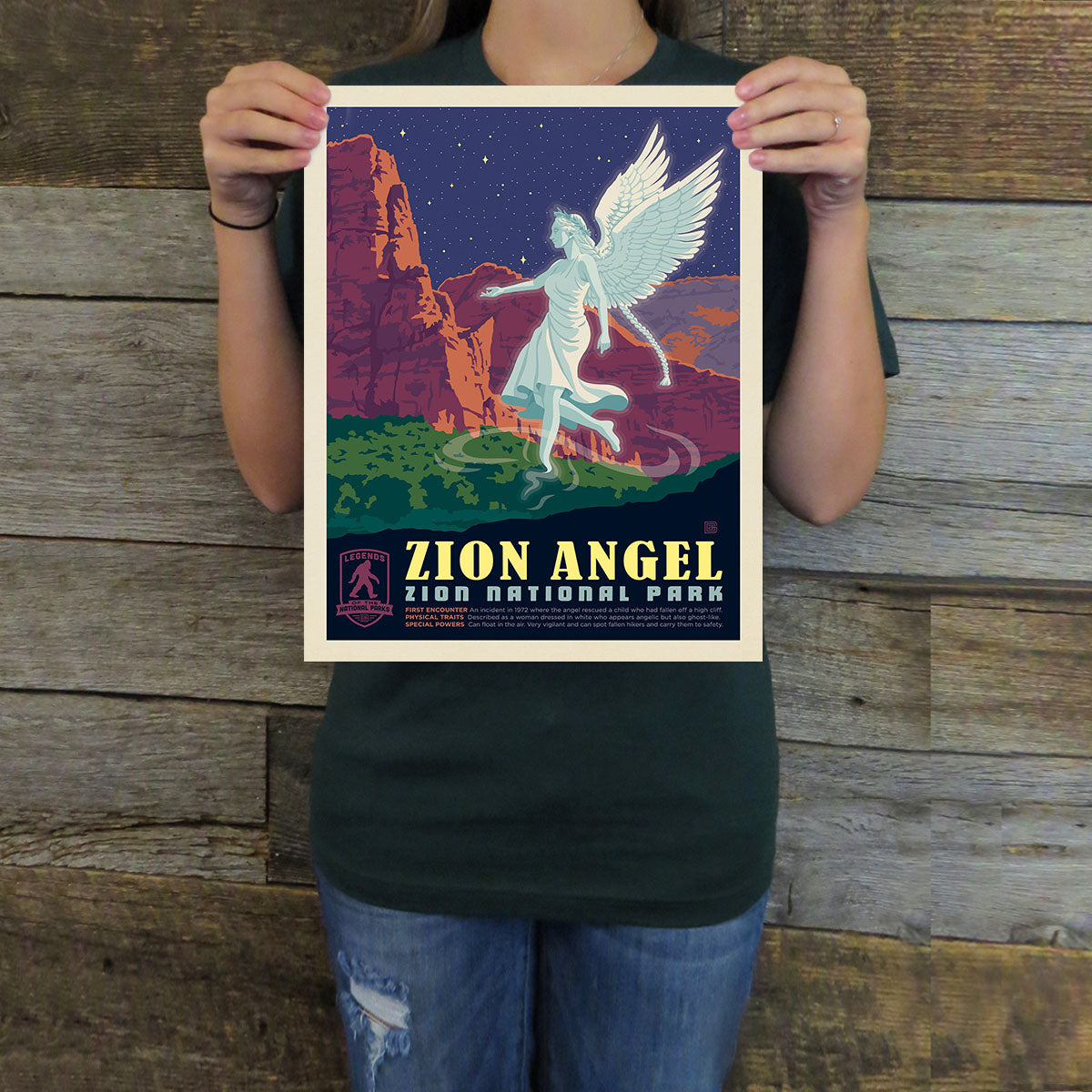 Person holding a Zion National Park poster with an angel design against a wooden background
