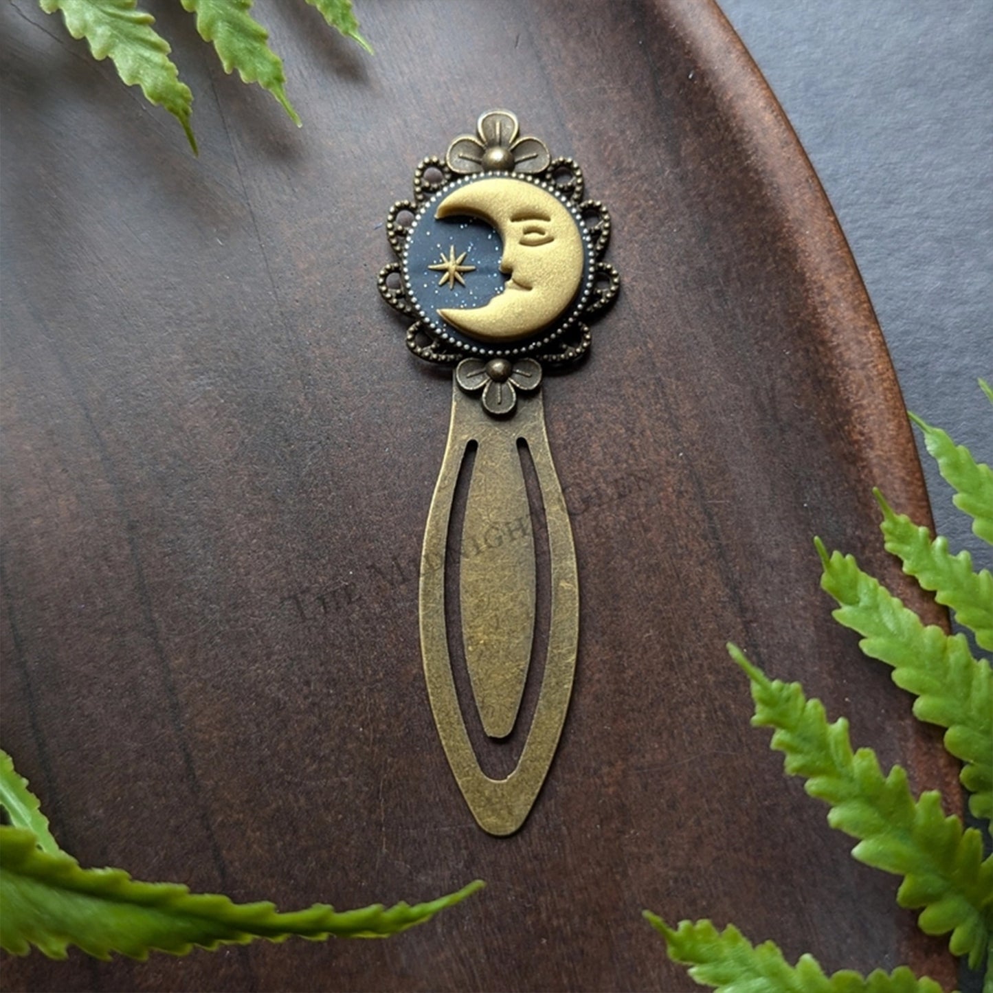 Decorative bookmark with moon and star design on a wooden surface with green leaves.
