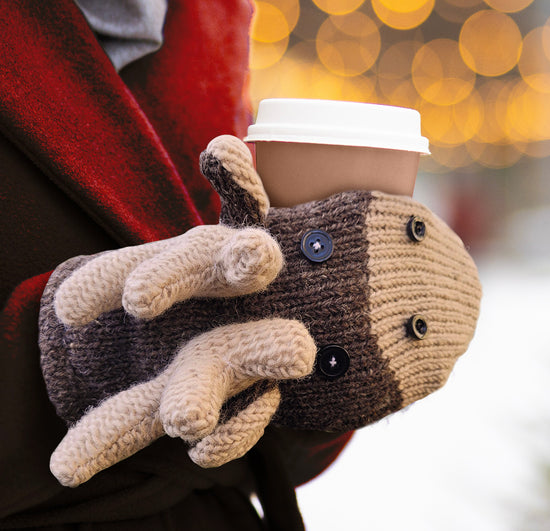 A model wearing a brown and tan mitten. The mitten is designed to look like moose heads, with black buttons for the eyes and nose, and woolen antlers sticking up at the middle. The model is holding a brown paper coffee cup in the mitten's mouth.