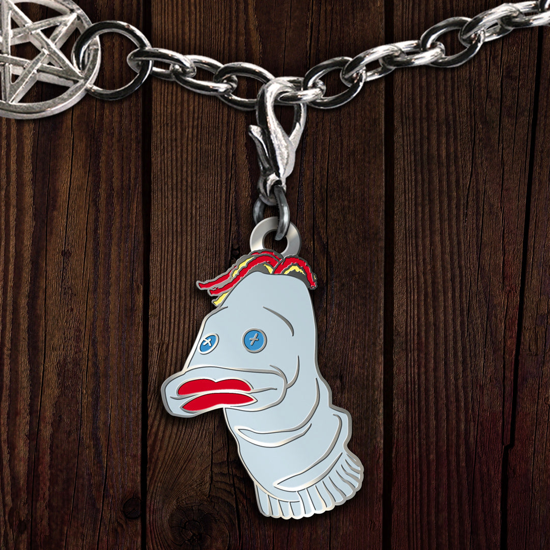 An enamel charm on a silver chain, against a wooden background. The charm depicts Mr. Fizzles, a sock puppet from the TV series "Supernatural." The puppet has blue buttons for eyes, red and yellow strings for hair, and bright red lips.