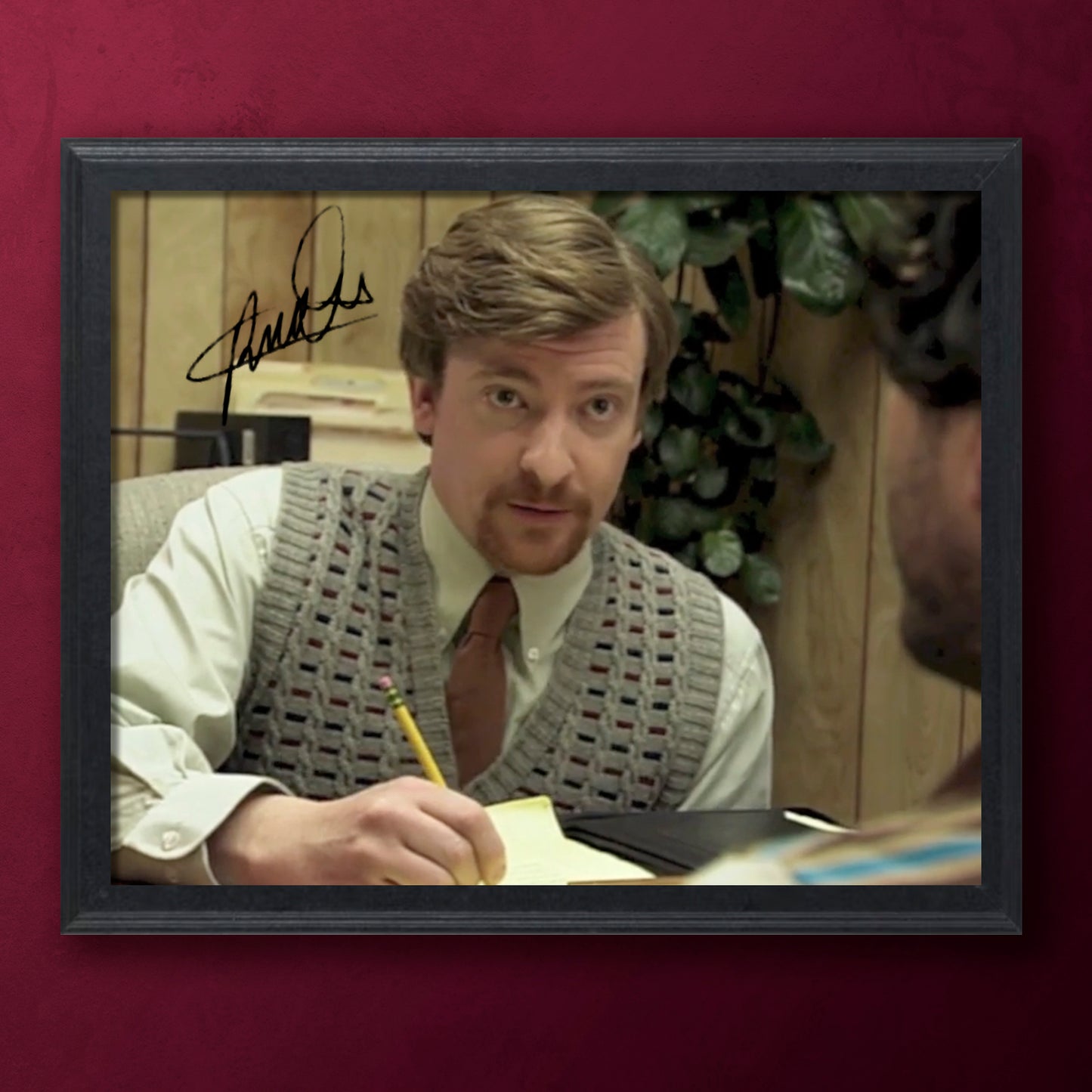 Framed photo of Rhys Darby in a gray vest holding a pencil, with a signature above his head on a red background.