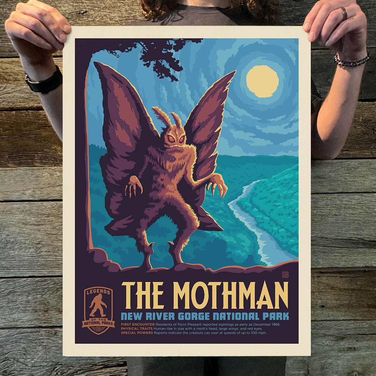 Poster of Mothman with a scenic background held by a person on a wooden surface
