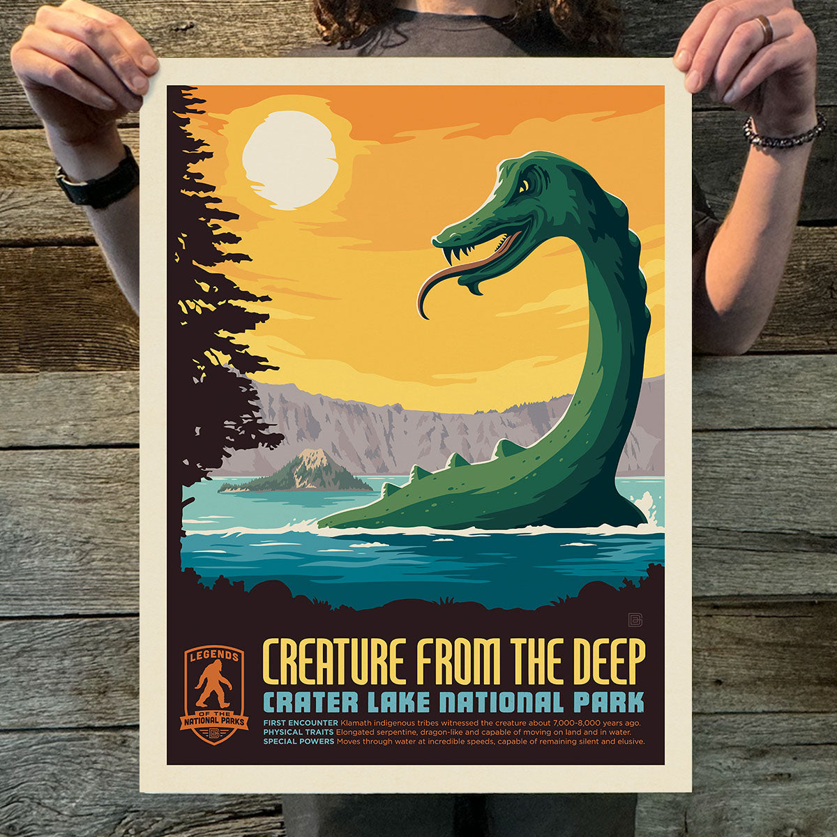 Poster of a creature from the deep at Crater Lake National Park held by a person on a wooden surface.