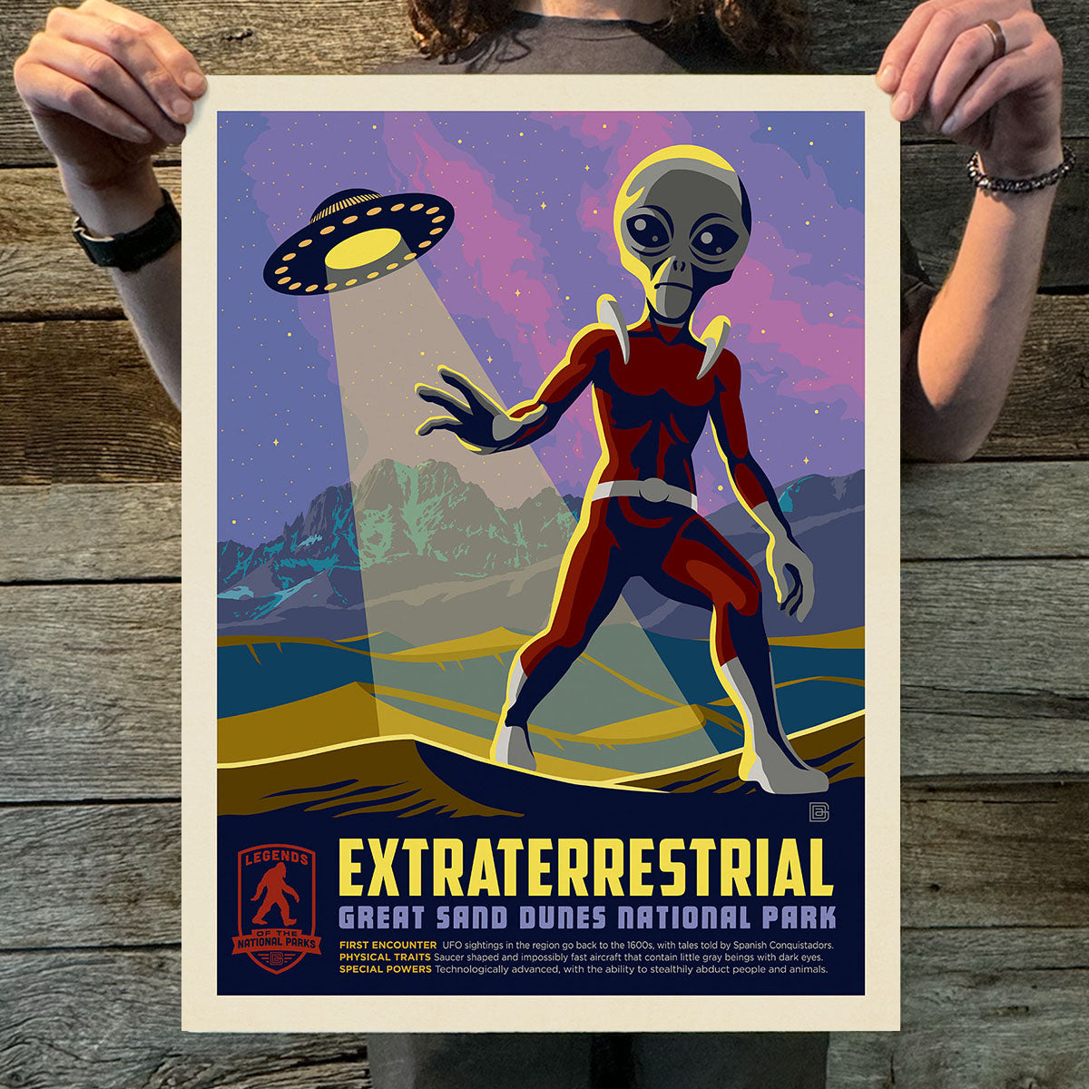 Poster of an alien in a red suit with a UFO and mountainous landscape, held by a person on a wooden surface.