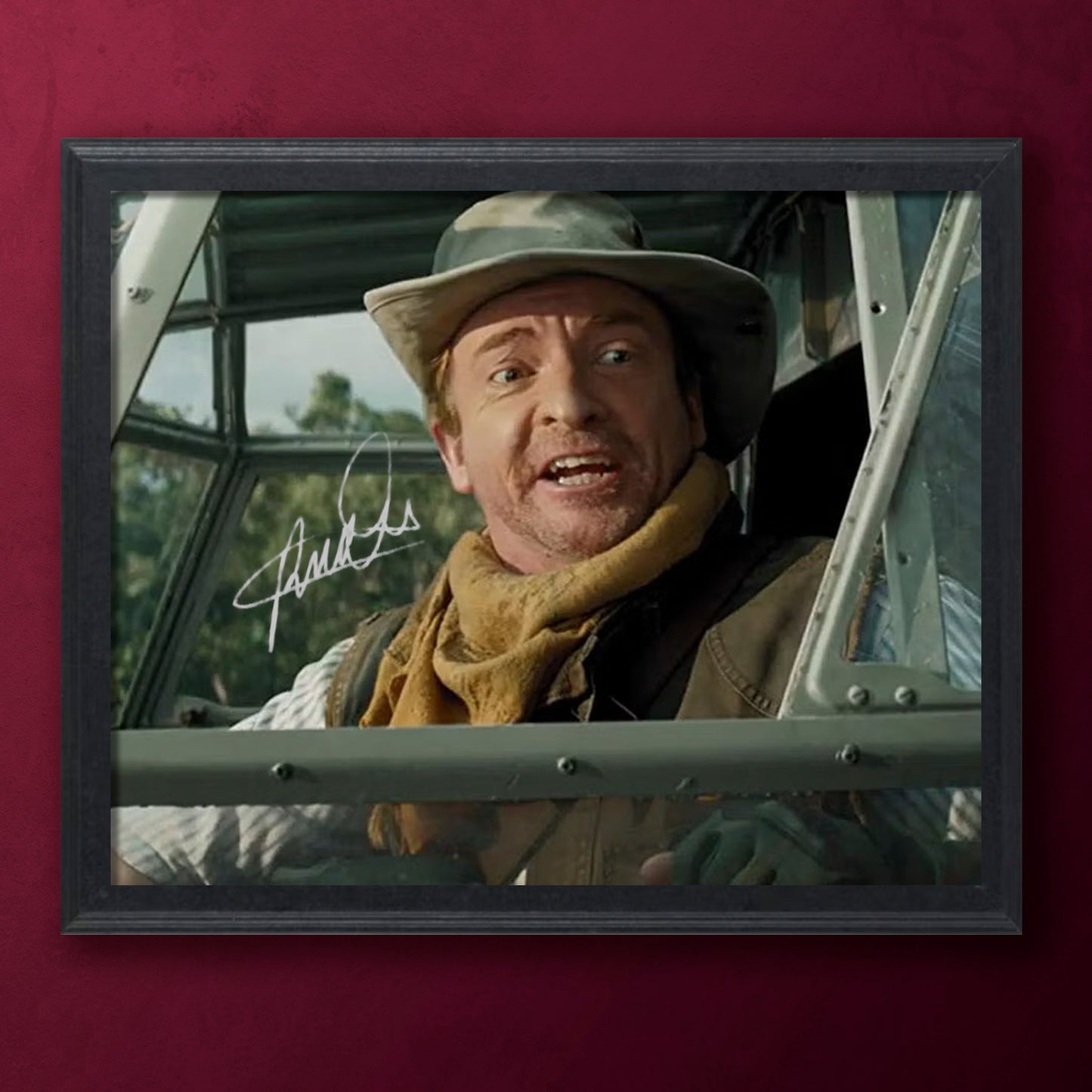 Framed photo of Rhys Darby wearing a hat and scarf, sitting inside a vehicle with a signature below.