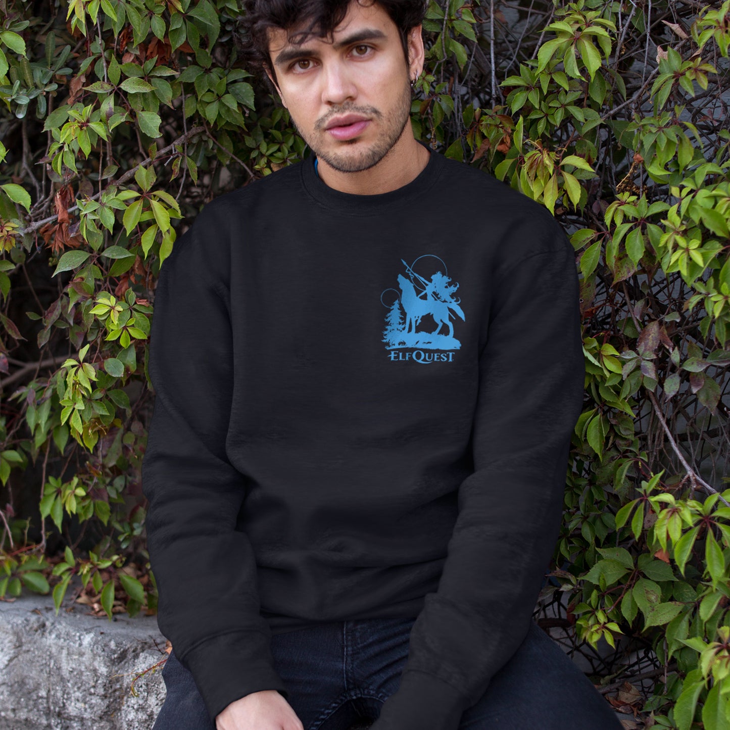 A male model in a black pullover sweatshirt. At the top  is a blue silhouette drawing of the character Nightfall, astride her wolf companion Spar. Nightfall is holding a spear. Behind her is a tree, with two moons in the sky. Underneath them is white text saying "ElfQuest."