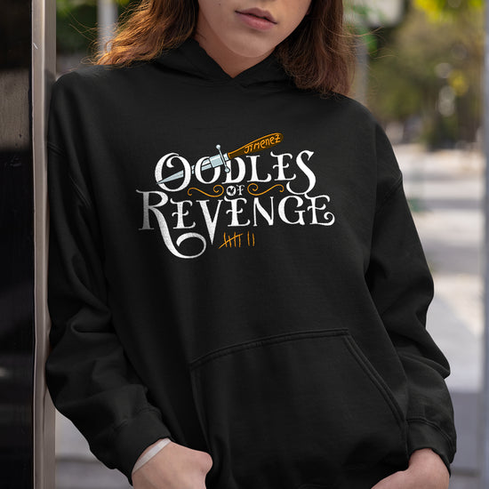 An image of a female model in a pullover hoodie, leaning against a wall. The front of the hoodie has white text that says "Oodle of revenge." Above the text is a drawing of a dagger, with the blade sticking through the openings of the two Os in oodles. Under the text are orange tally marks, counting to 7. Behind the model is a street lined with trees.