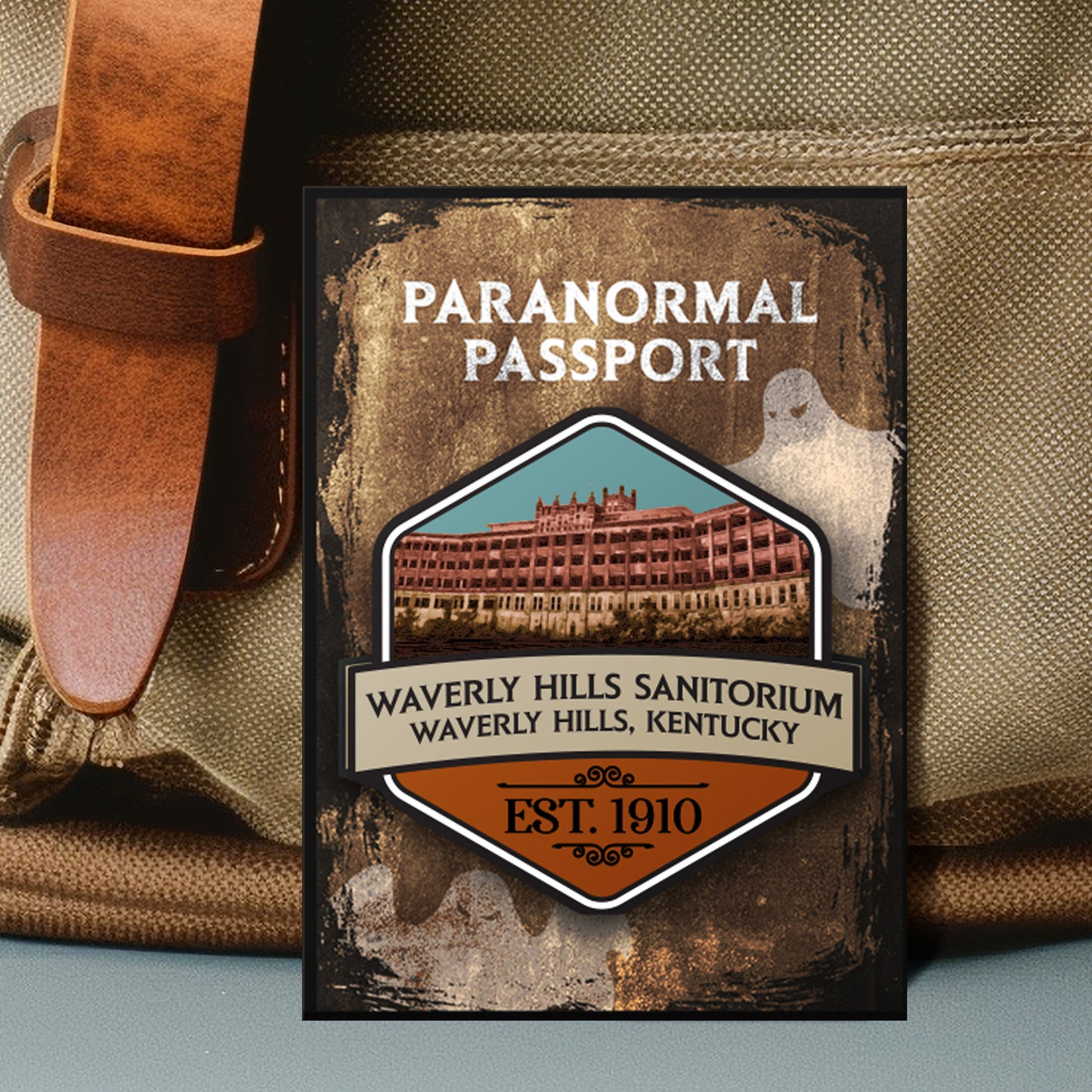 Close up on an enamel pin. The pin has a drawing of a run down sanitorium in the center, surrounded by a stone-colored background. Two white ghost drawings are in the background. At the top is white text saying "Paranormal passport." Under the battlefield drawing is text saying "Waverly Hills Sanitorium. Waverly Hills, Kentucky. 1910."