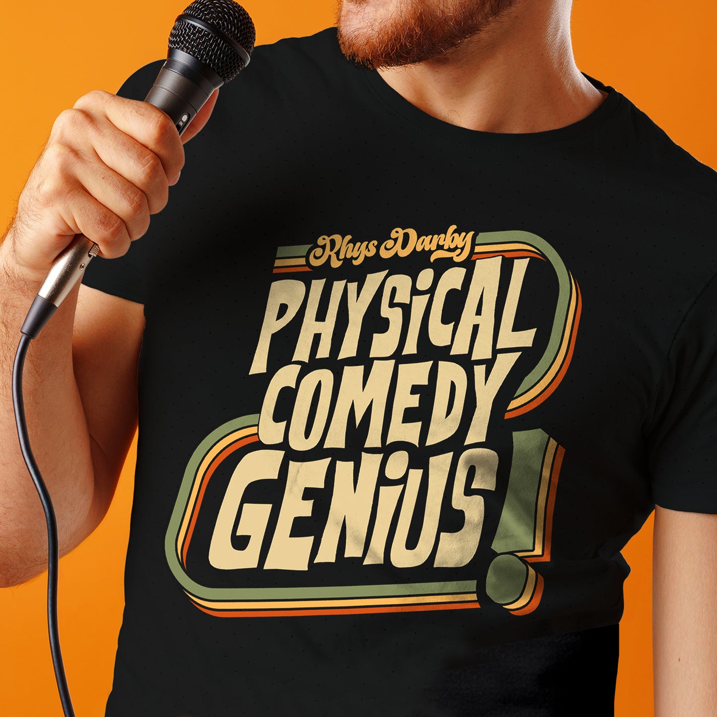 Person wearing a black t-shirt with 'Physical Comedy Genius' text, holding a microphone against an orange background