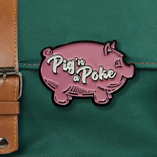 A pink pig-shaped enamel pin on a green canvas messenger bag. Across the middle of the pig is white scripted text that says "pig n a poke. A brown leather bag closure is next to the pin.