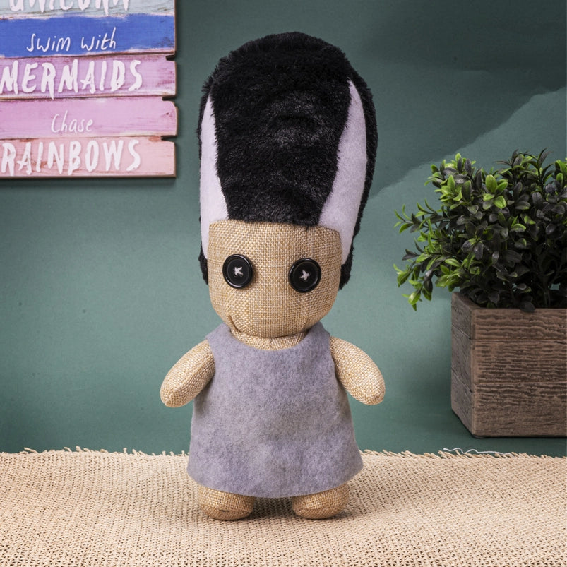 Plush toy with black and white hair and a gray dress on a textured surface with a plant and colorful wall in the background.