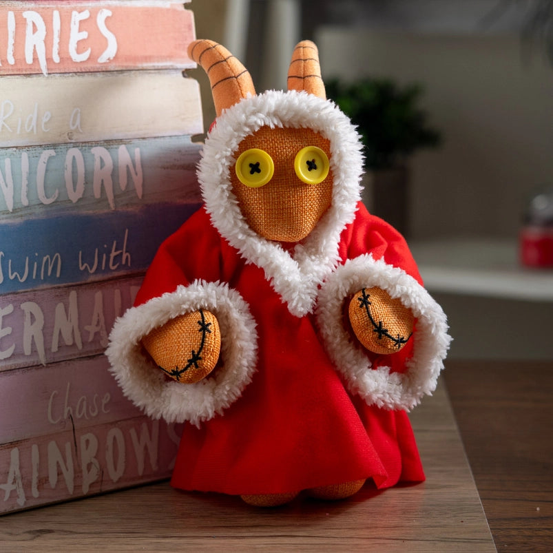 A stuffed plushie on a wooden table, leaning against a rainbow colored wooden sign. The plushie has yellow buttons as its eyes, and curled horns at the top of its head. It is wearing a red robe with white edges.