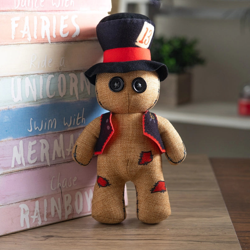 A stuffed plushie on a wooden table, leaning against a rainbow colored wooden sign. The plushie has a black and red top hat and vest, with black buttons as eyes. A patch on its hat has the number 13 in red.