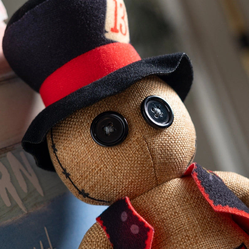 Close up of a stuffed plushie's face. The plushie has black button eyes, and is wearing a red and black vest and top hat. The hat has a white patch with a red 13 on it.