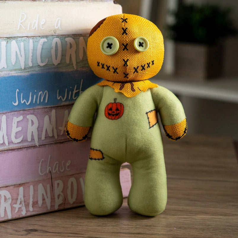 A stuffed plushie on a wooden table, leaning against a rainbow colored wooden sign. The plushie has light green buttons as its eyes, and is wearing a green jumpsuit with an orange sack as its head.