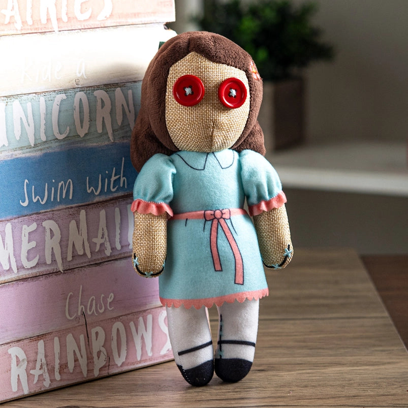 A stuffed plushie on a wooden table, leaning against a rainbow colored wooden sign. The plushie depicts one of the twin girls from the movie "The shining," and is wearing a blue dress with pink frills and a belt. The plushie has two red buttons as its eyes.