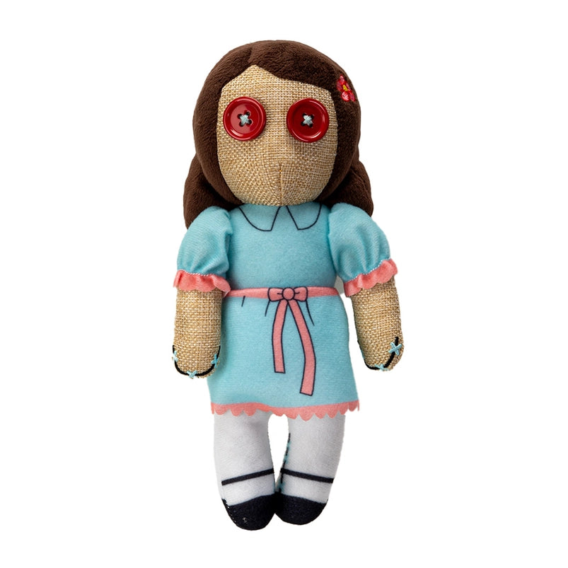 A stuffed plushie on a wooden table, against a white background. The plushie depicts one of the twin girls from the movie "The shining," and is wearing a blue dress with pink frills and a belt. The plushie has two red buttons as its eyes.