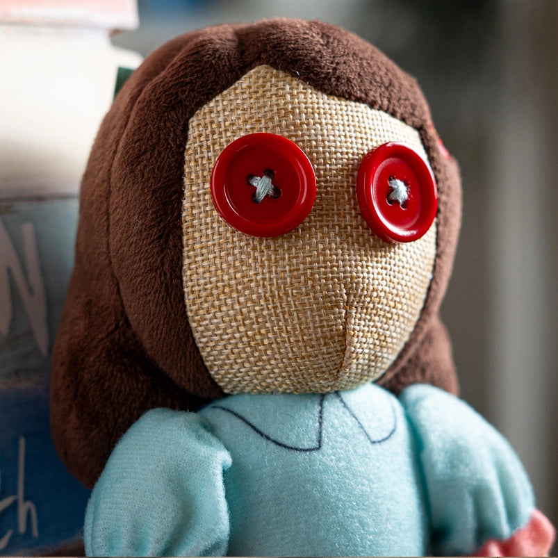 Close up of a stuffed plushie's face. The plushie has red buttons as its eyes, and is wearing a blue dress with pink frills.
