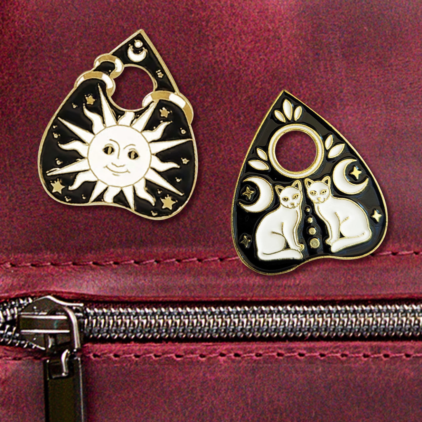 A close-up photo of two black-and-gold planchette pins on a maroon leather purse. The left pin depicts a large white sun in a tarot-like art style, surrounded by small gold stars. The right pin depicts two identical white cats facing each other, each with a moon and two small stars above/around them.