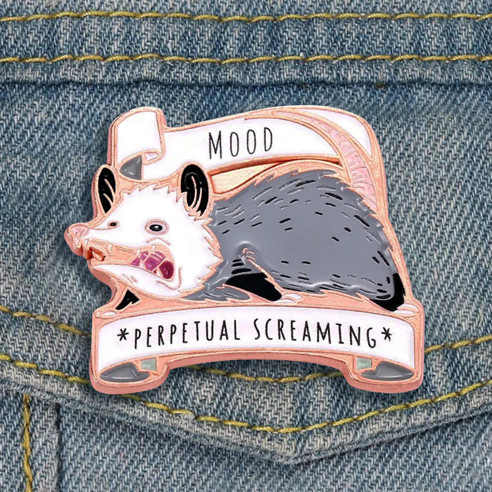 Close up view of an enamel pin on a light demin lapel. The pin depicts a cartoonish opossum, mouth wide open in a scream. Above and below the opossum are white ribbons with black text saying "Mood: perpetual screaming."