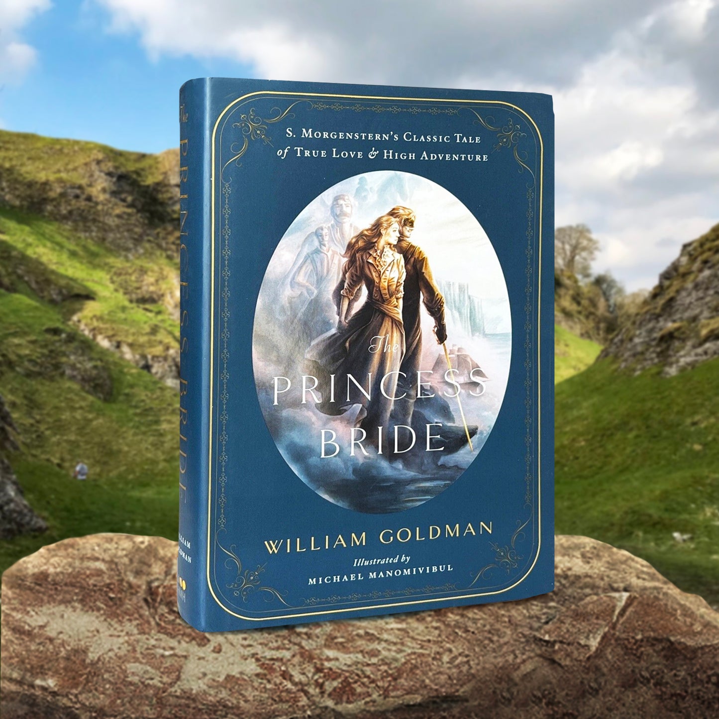 A blue book on a brown rock, with hills in the background. White text on the front says "The Princess Bride" by William Goldman, reading "S. Morgenstern's Classic Tale of True Love & High Adventure - Illustrated by Michael Manomivibul" In the center is a drawing of characters from the book.