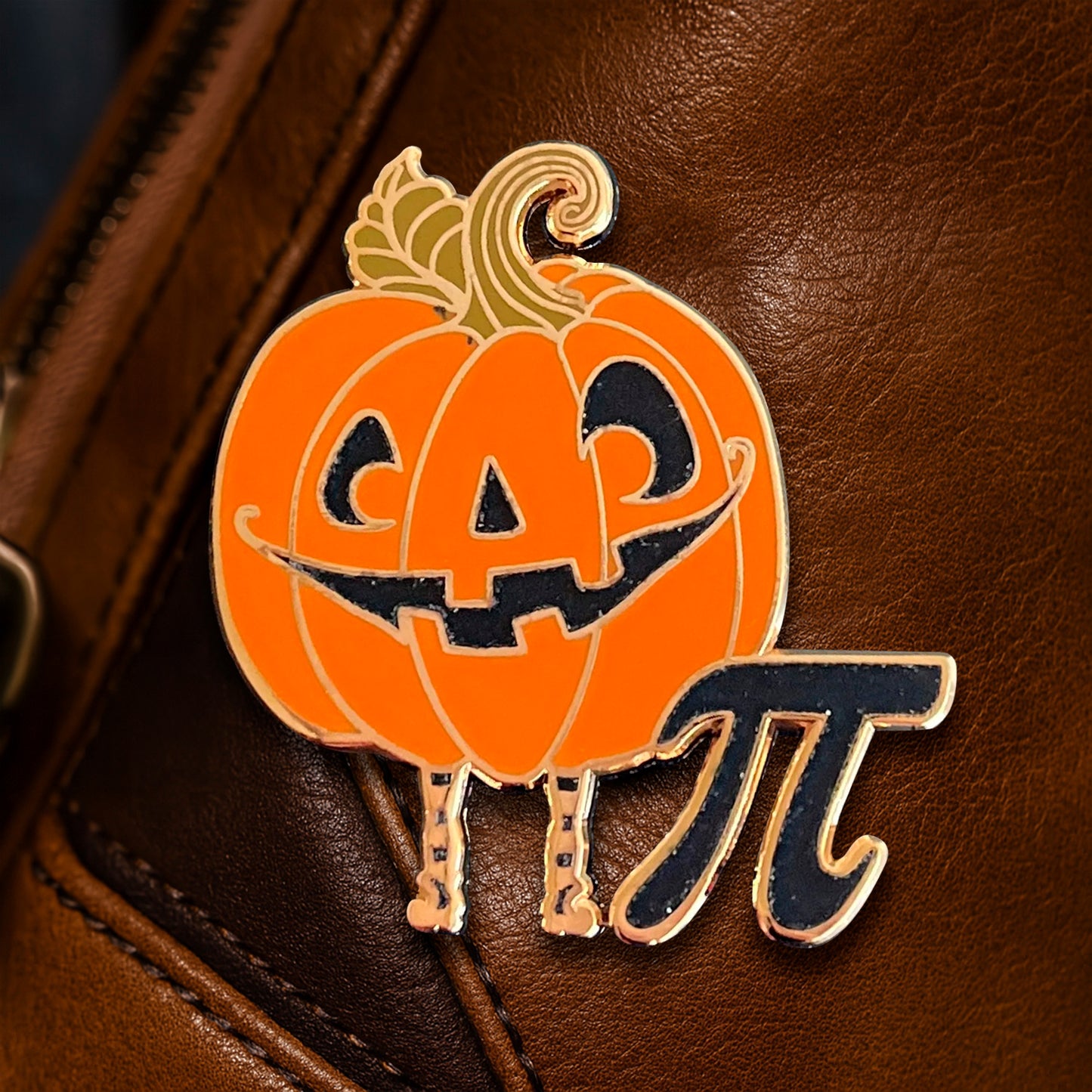 A Halloween-themed enamel pin shaped like a smiling orange jack-o'-lantern with a gold stem and legs, next to a navy blue pi symbol with a gold outline, displayed on a brown leather surface.
