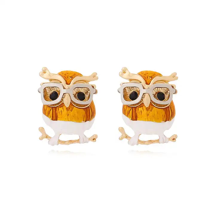 Close up view of a pair of earrings against a white background. Each earring is shaped like a small brown owl perched on a brass tree branch. The owls are white from the middle down, and are wearing white glasses.