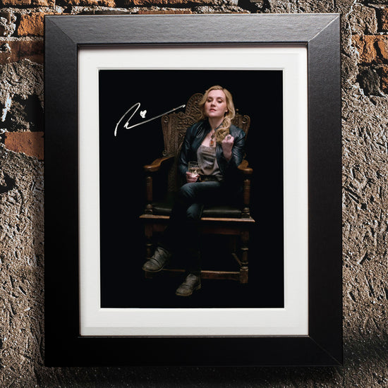 A black framed photograph against a concrete and brick wall. The photograph depicts actor Rachel Miner, dressed as the character Meg from the series "Supernatural." Rachel is sitting on a wood chair with an ornately carved background. She is holding a glass in one hand, and checking the fingernails on her other hand. In the top left corner is Rachel's autograph in white ink.