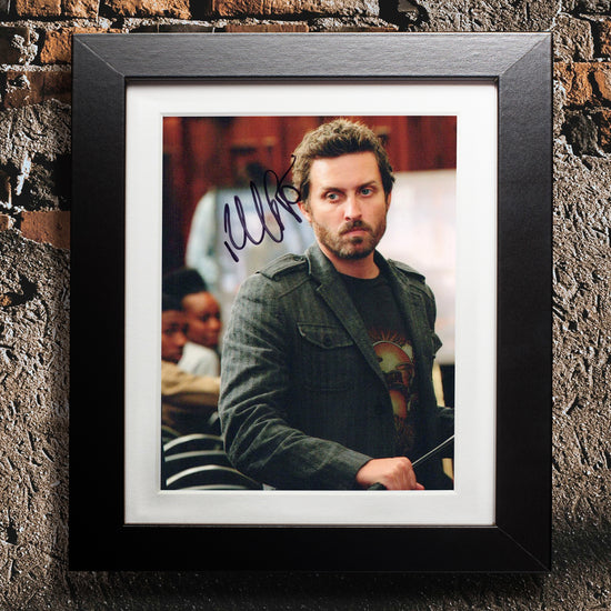 A black framed photograph against a brick and concrete wall. The photograph depicts actor Rob Benedict, dressed as Chuck Shurely from the series "Supernatural." Rob is in a black tee shirt and green jacket, holding a metal bar in his hands. On the top left side is Rob's autograph in black ink.