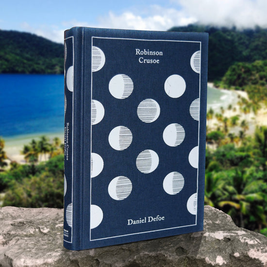 A dark blue book with white circles across the cover, standing on a rock ledge. White text says "Robinson Crusoe, Daniel Defoe." In the background is a beach on a tropical island, surround by trees and blue water.