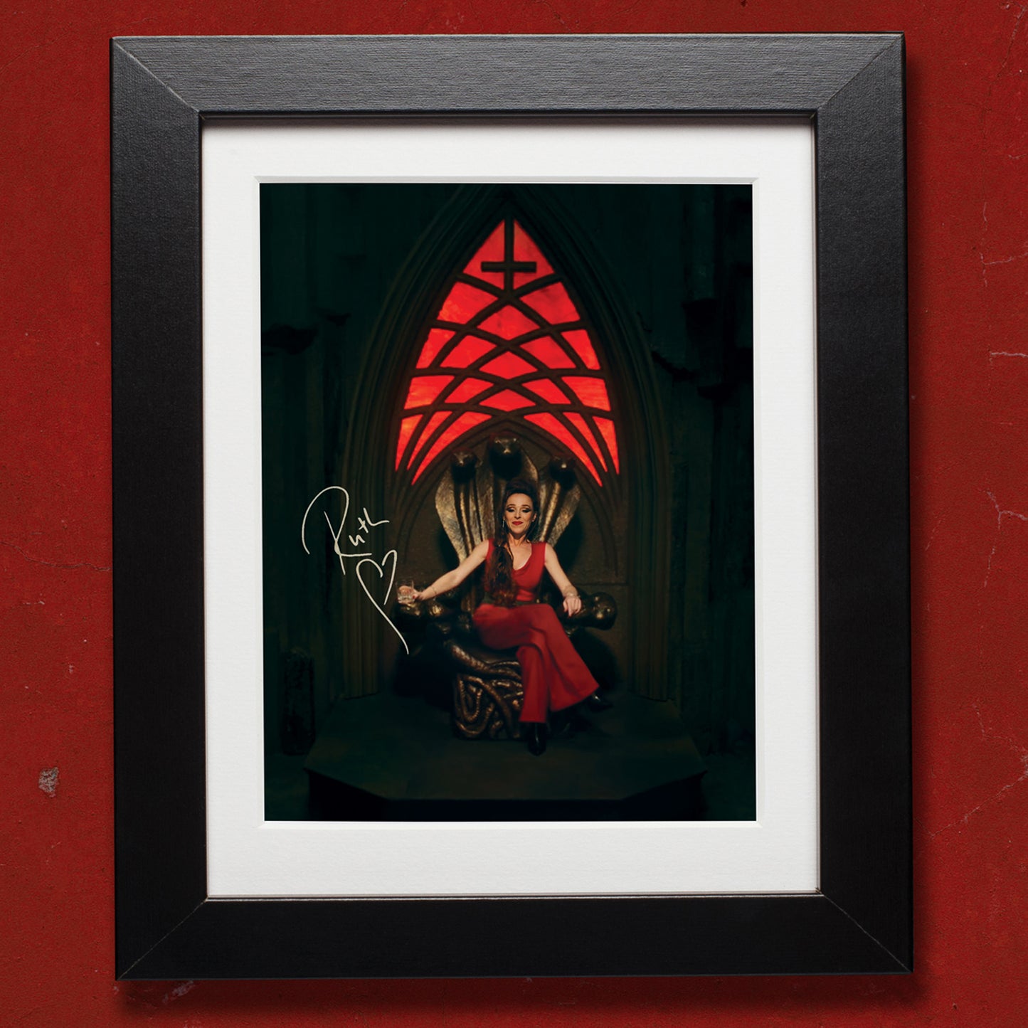 A picture in a black frame against a dark red wall. The picture is of Ruth Connell as the character "Rowena" from the TV series Supernatural, sitting on her throne. Above her is a red stained glass window with an upside down cross at the top. Next to her is Ruth's signature in white ink.