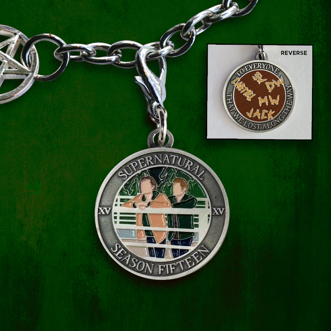 Front and back views of a bracelet charm. On the front is a depiction of Sam and Dean Winchester leaning on a white fence, with trees in the background. Around the edge of the coin, raised text says "Supernatural Season Fifteen.” The back of the charm depicts a wooden table with the names and initials of SPN characters carved into it. Around the edge is raised text saying “To everyone that we lost along the way.”