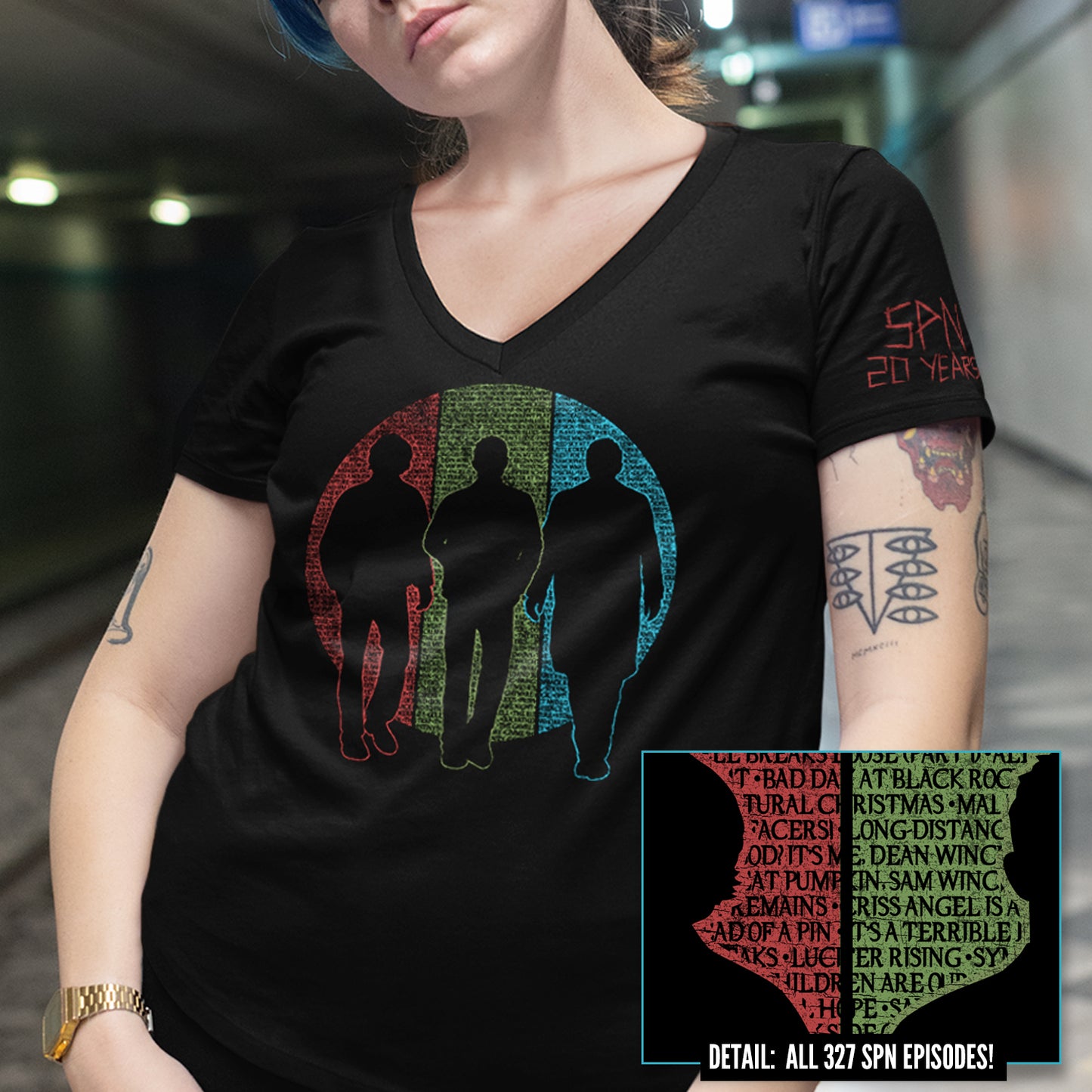 A female model wearing a black v-neck shirt. The shirt features silhouettes of Sam and Dean Winchester and Castiel. Behind them is a circle split into vertical thirds - a red slice behind Sam, green behind Dean, and blue behind Castiel. The circle is filled with the titles of all 327 episodes of Supernatural in black font. One short sleeve reads 'SPN 20 YEARS' in a capital font with a 'carved' effect.