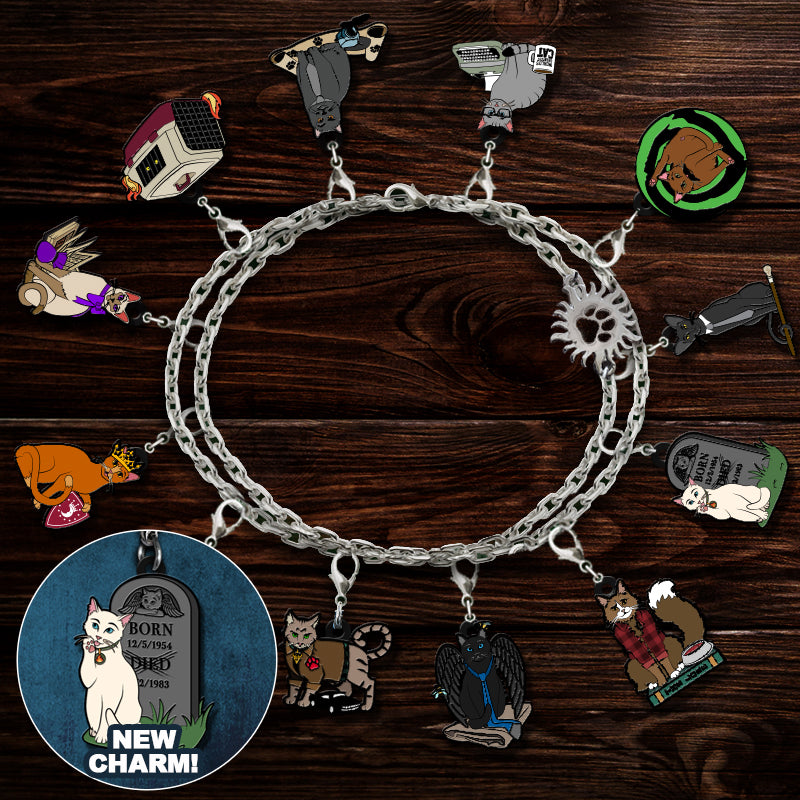 A silver-tone chain bracelet with multiple cat-themed charms, displayed on a dark wood background. The charms include a tombstone, a cat in a bed, a cat wearing sunglasses, a cat in a plaid shirt, a bird, a cat on a skateboard, a cat in a wheelchair, a cat wearing a hat, a cat in a green outfit, and a round badge that says 'BORN TO BE A CAT' and 'NEW CHARM!'.