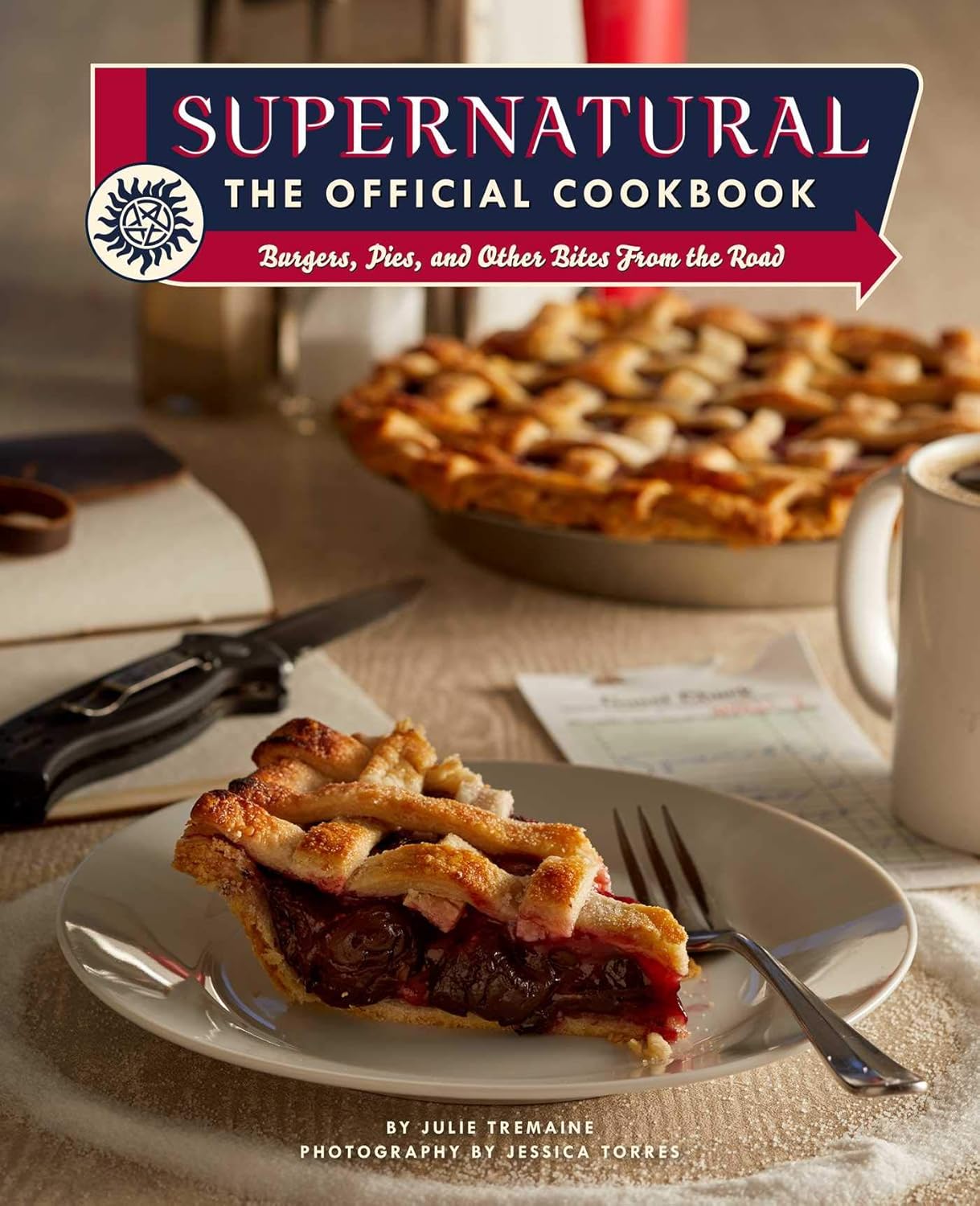 A hardcover cookbook on a white background. The cookbook has a picture of a slice of pie on a plate, with "supernatural, the official cookbook" at the top in white letters.
