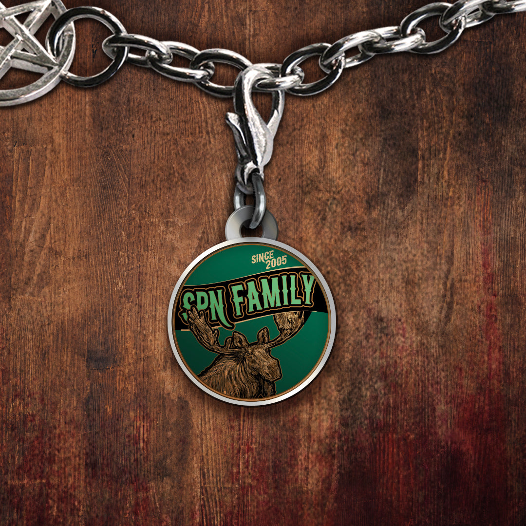 A silver-toned charm featuring a moose illustration and the text 'SPN FAMILY' on a dark green background, displayed against a rustic brown wood surface.