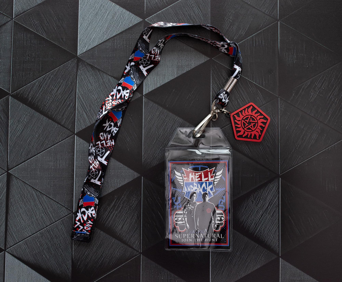 A lanyard with an anti-possession charm in red and black attached to the end. The lanyard features "to hell and back" in white text. Next to the lanyard is a badge featuring Sam and Dean Winchester. A red and blue road symbol at the top has white text saying to hell and back. The lanyard and badge are on a black surface.