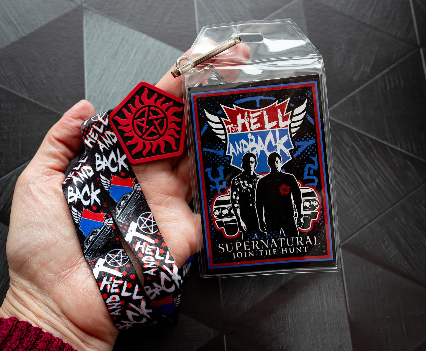 A model's hand holding a lanyard with an anti-possession charm in red and black attached to the end. The lanyard features "to hell and back" in white text. Next to the lanyard is a badge featuring Sam and Dean Winchester. A red and blue road symbol at the top has white text saying to hell and back. The lanyard and badge are above a black surface.