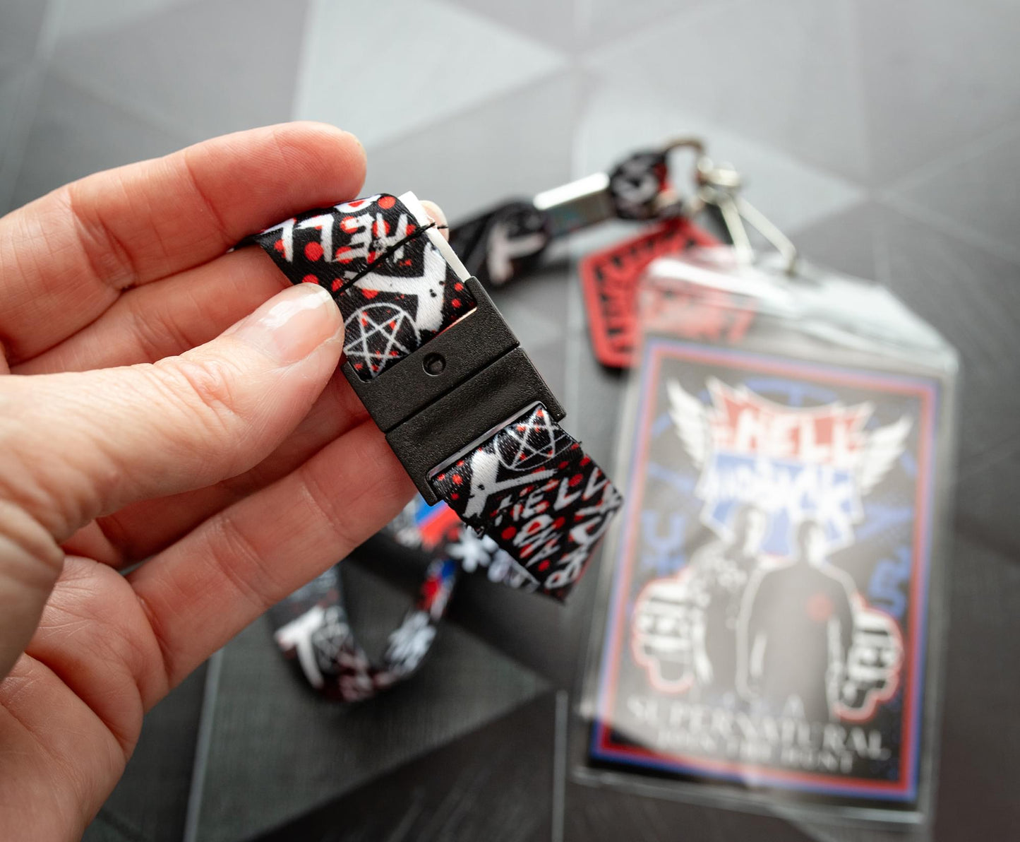 A model's hand holding a lanyard with an anti-possession charm in red and black attached to the end. The lanyard features "to hell and back" in white text. Next to the lanyard is a badge featuring Sam and Dean Winchester. A red and blue road symbol at the top has white text saying to hell and back. The lanyard and badge are above a black surface.