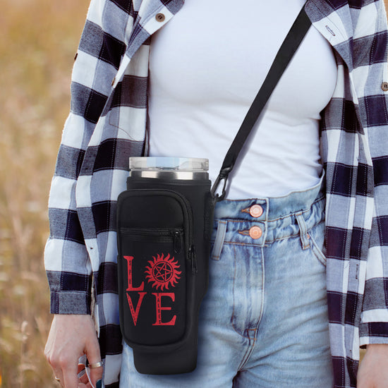 A female model wearing a black and white plaid shirt over a white t-shirt. Slung over her shoulder is a black caddy bag, hanging at her hip level. The front of the bag has red text saying Love, with the "o" replaced by the anti-possession symbol. The bag has several zippered pockets, and a large travel mug is visible in the bag's top pocket. Behind the model is a field. 